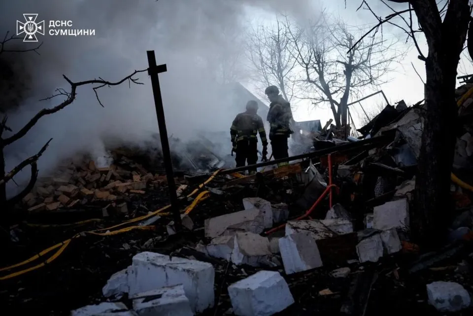 Două persoane în uniforme stau printre dărâmături și resturi de zidărie sub un cer plin de fum și copaci fără frunze.
