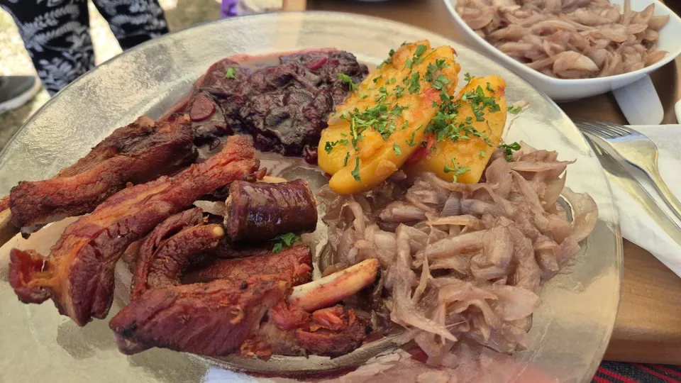 O farfurie mare cu carne de porc friptă, costuri, zăcări de cartofi galbeni ornate cu pătrățele verzi, vară roșie și ceapă roșie tocată fin.