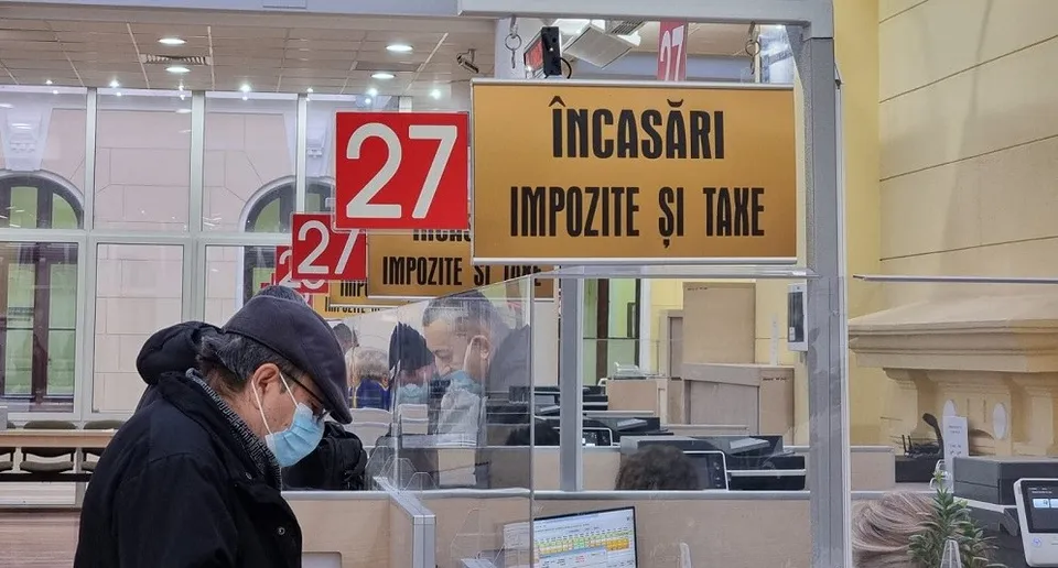 Un bărbat cu șapcă și mască medicală stă în fața unui ghișeu de încasări marcat cu numărul 27 și textul impozite și taxe.