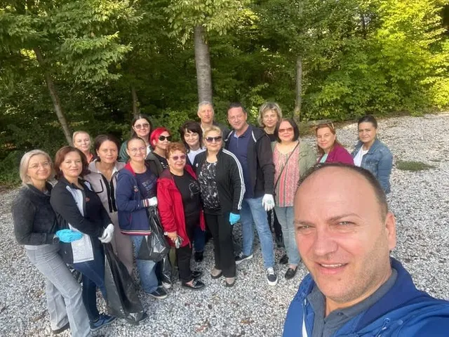 Un grup de persoane stă în picioare pe un drum de pietriș, în fața unor copaci verzi, purtând haine casual și mănuși de protecție.