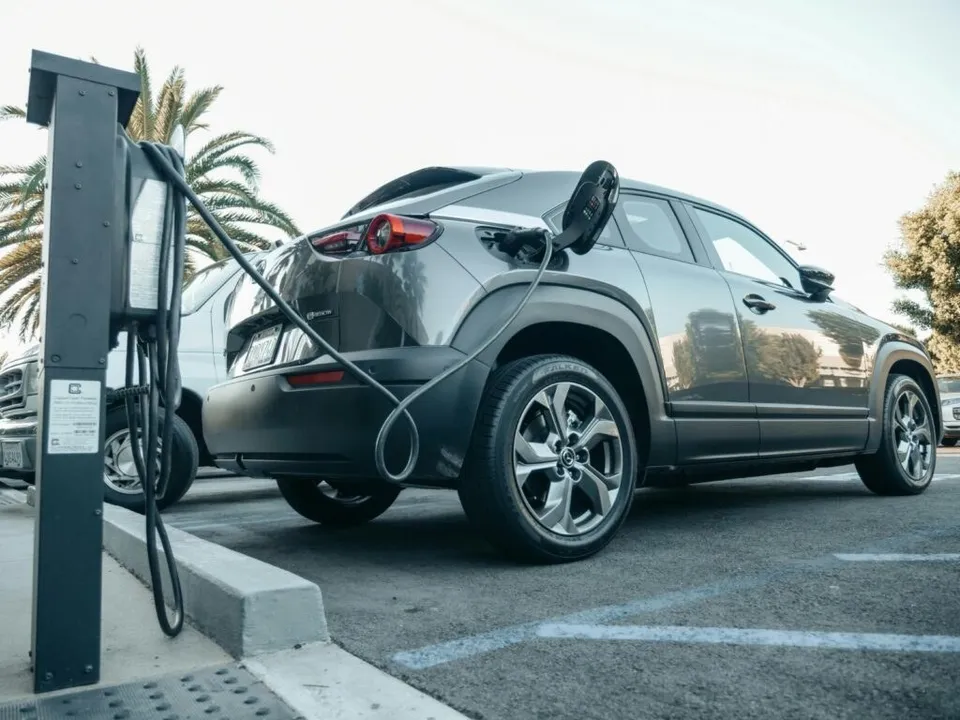 Un autoturism gri este conectat la o stație de încărcare electrică într-o parcare cu palmieri în fundal.