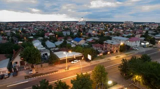 O vedere panoramică asupra unui oraș cu numeroase case cu acoperișuri colorate, străzi iluminate și clădiri dispersate sub un cer înnorat la amurg.
