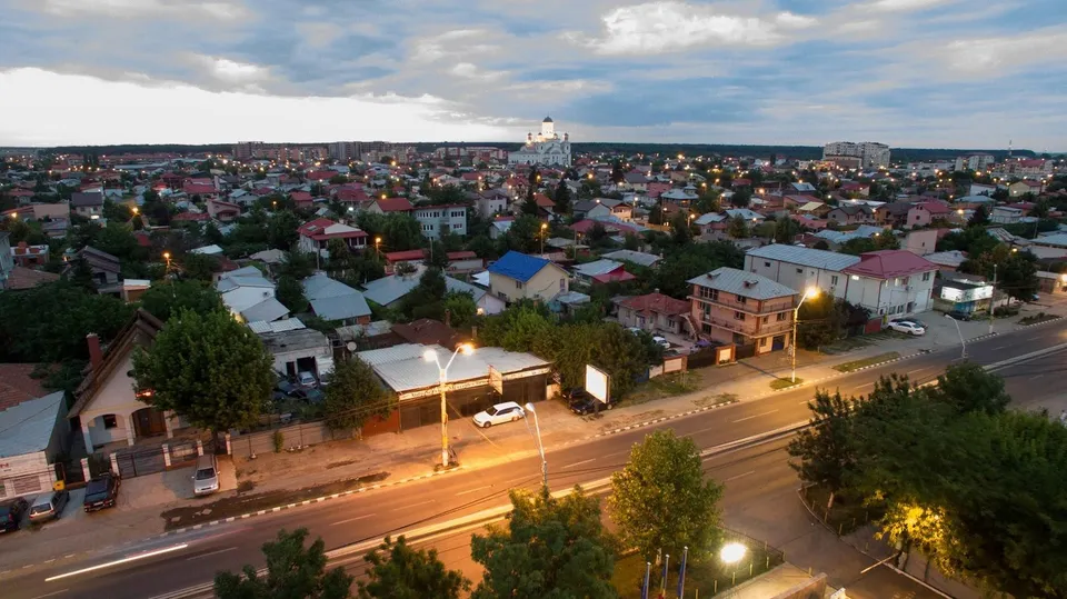 O vedere panoramică asupra unui oraș cu numeroase case cu acoperișuri colorate, străzi iluminate și clădiri dispersate sub un cer înnorat la amurg.