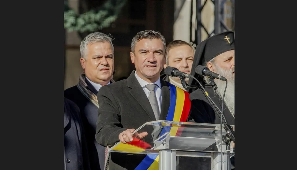 Un bărbat într-un costum gri și cravată deschide un podium transparent, purtând o eșarfă tricoloră, în fața altor trei bărbați.