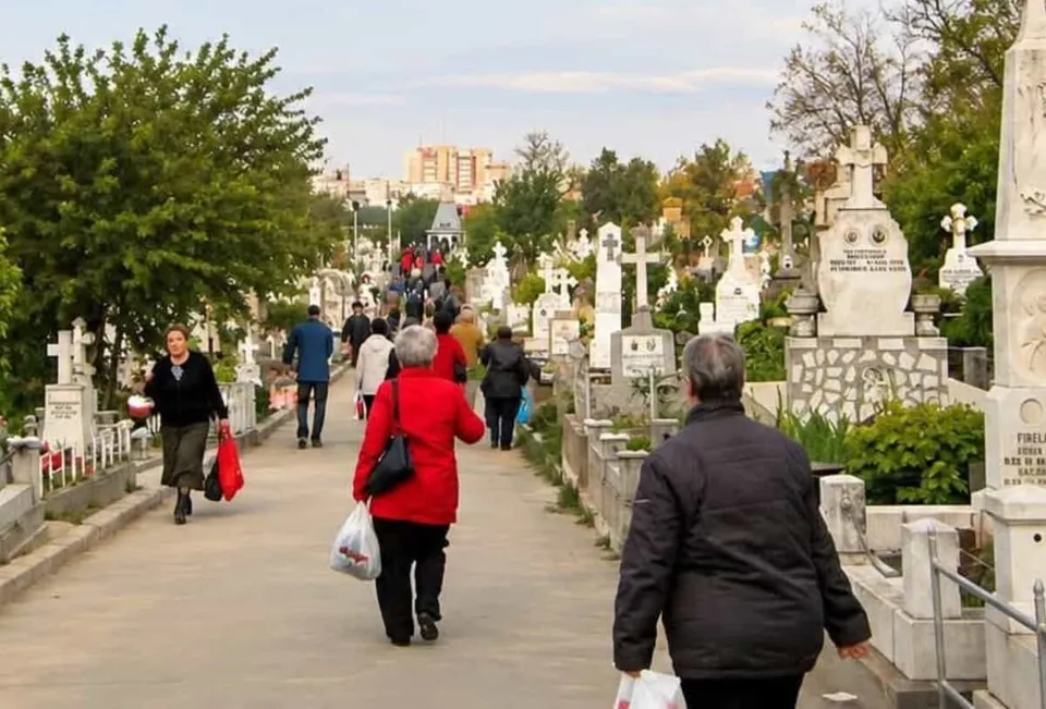 Oamenii merg pe un drum pavat printre cruci de piatră și monumente funerare într-un cimitir, sub un cer înnorat.