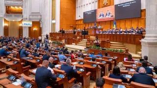 Oamenii stau la mese de lucru în fața unui podium înalt într-o sală de conferințe mare cu panouri de lemn și ecrane digitale.