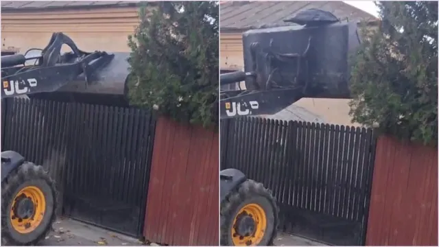 Un excavator cu brandul JCB ridică un sac negru de deșeuri deasupra unui gard metalic negru și al unui gard maroniu.