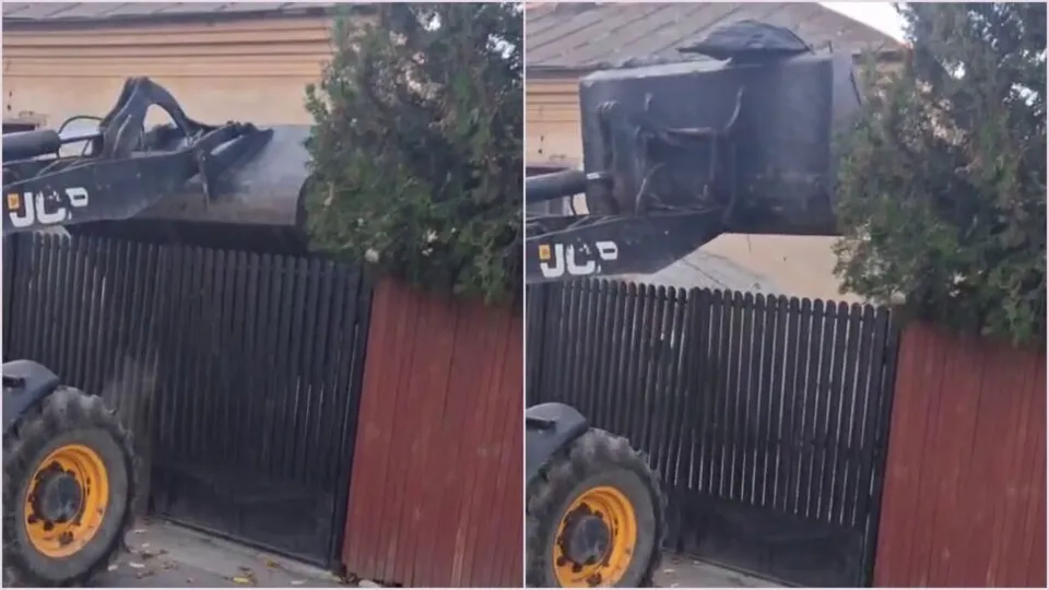 Un excavator cu brandul JCB ridică un sac negru de deșeuri deasupra unui gard metalic negru și al unui gard maroniu.