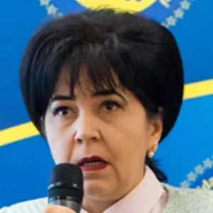O femeie cu părul negru și buze roșii vorbește într-un microfon în fața unui fundal albastru cu elemente galbene.
