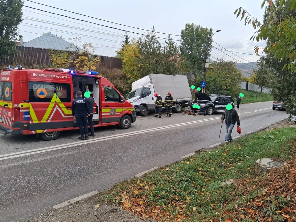 O ambulanță roșie și galbenă stă pe marginea drumului lângă un vehicul alb și o mașină neagră, în timp ce mai mulți oameni stau pe carosabil.