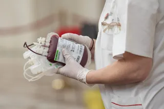 O persoană îmbrăcată în halat alb și mănuși medicale ține un sac de colectare a sângelui cu etichetă albă.