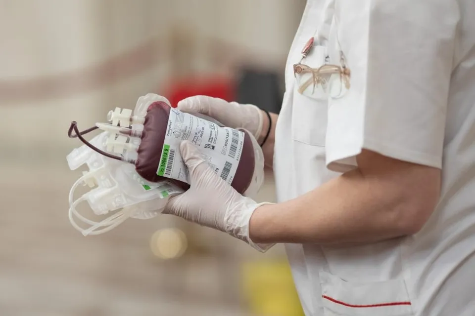 O persoană îmbrăcată în halat alb și mănuși medicale ține un sac de colectare a sângelui cu etichetă albă.