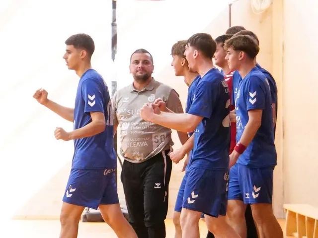 Câțiva tineri sportivi în echipamente albastre de handbal stau lângă un bărbat cu tricou gri într-o sală de sport.