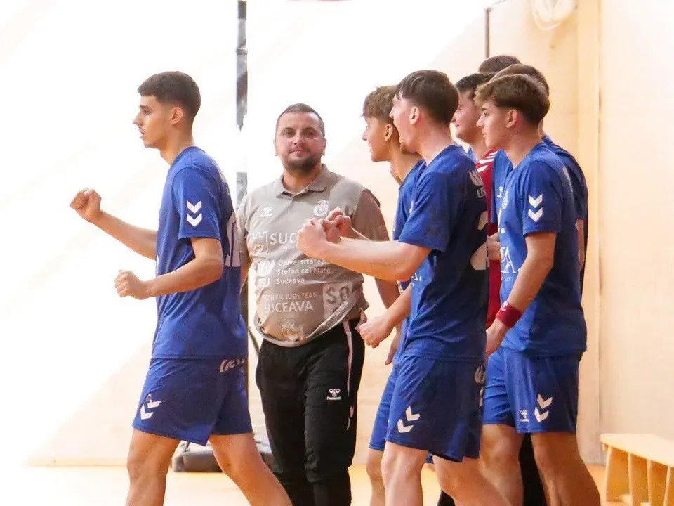 Câțiva tineri sportivi în echipamente albastre de handbal stau lângă un bărbat cu tricou gri într-o sală de sport.