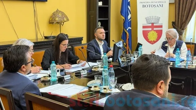 Oameni stau la o masă lungă cu documente, apă și microfoane într-o încăpere cu pereți galbeni și un steag.