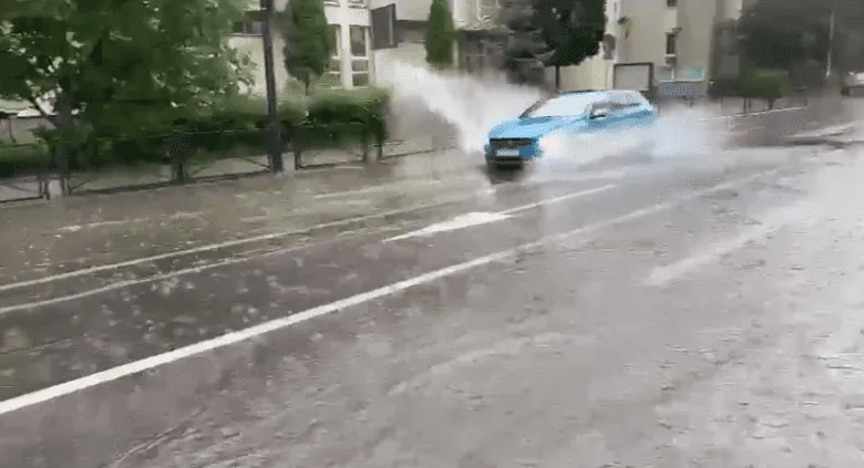 Un autovehicul albastru circulă pe un drum umed, ridicând o spray de apă din cauza unei bălți.