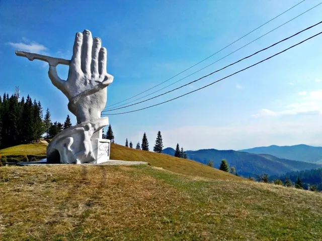 O sculptură gri în formă de mână cu degete lungi este situată pe un deal de iarbă uscată sub un cer senin.