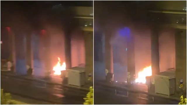 Flăcări mari și fum se ridică de la un obiect de pe un trotuar în fața unei clădiri pe timp de noapte.