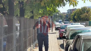 Un bărbat cu o cămașă închisă la culoare și pantaloni închiși merge pe o trotuar lângă mașini parcate și un gard metalic.