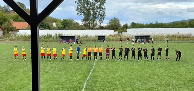 Oamenii de pe teren de fotbal sunt așezați în două rânduri pe iarbă verde, purtând echipamente sportive de culori galben, roșu sau negru.