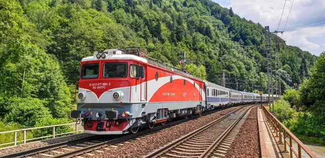 Un tren feroviar roșu și alb cu vagoane albastre circulă pe o cale ferată lângă un versant de munte acoperit de vegetație verde densă.