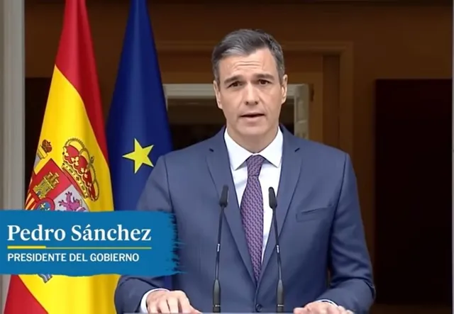 Un bărbat cu părul scurt, îmbrăcat în costum albastru, cămașă albă și cravată mov, stă în fața unui podium cu microfoane și a două steaguri.