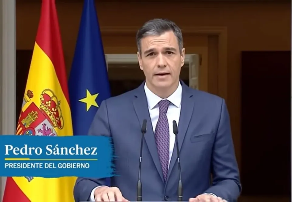 Un bărbat cu părul scurt, îmbrăcat în costum albastru, cămașă albă și cravată mov, stă în fața unui podium cu microfoane și a două steaguri.