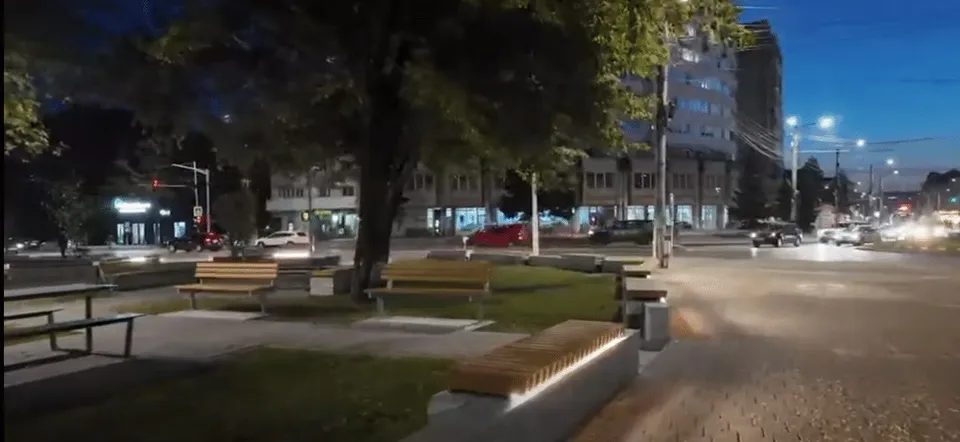 O zonă de odihnă nocturnă prezintă bănci de lemn, pavaj, spații verzi și clădiri urbane sub un cer închis.