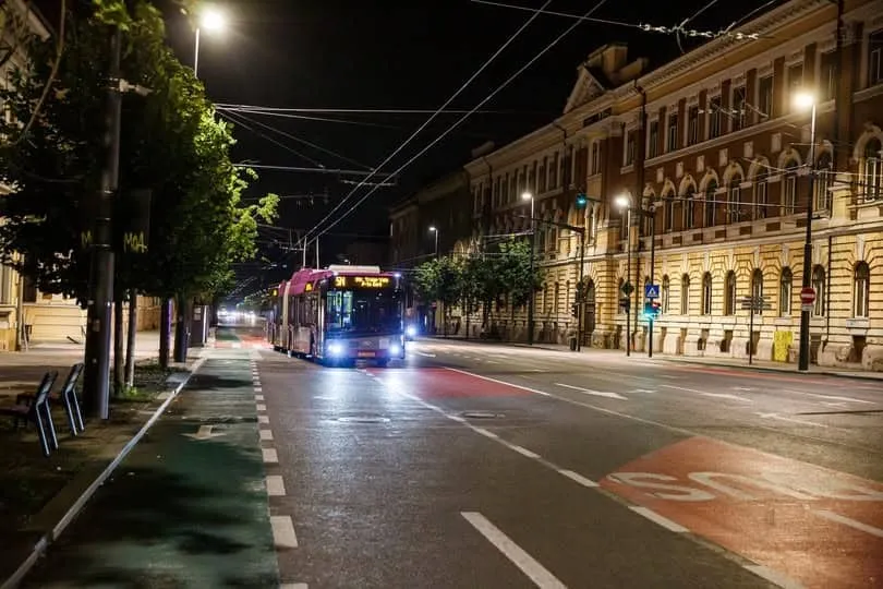 Un autobuz alb și albastru circulă pe o stradă asfaltată, lângă o pistă pentru biciclete, sub luminile străzii pe timp de noapte.