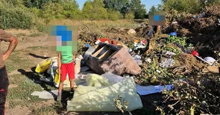 O persoană în bermudaج roșii și tricou verde stă lângă un morman de deșeuri compus din obiecte diverse, materiale de construcții și resturi vegetale.