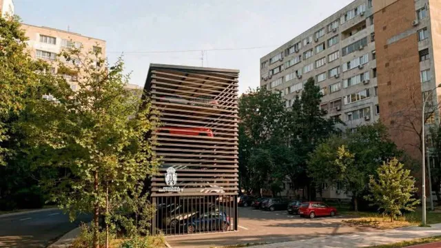 O structură metalică cu rafturi orizontale pentru parcări este situată între copaci și clădiri de locuințe înalte.