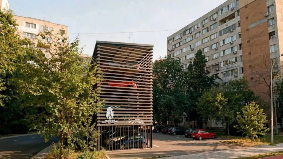 O structură metalică cu rafturi orizontale pentru parcări este situată între copaci și clădiri de locuințe înalte.