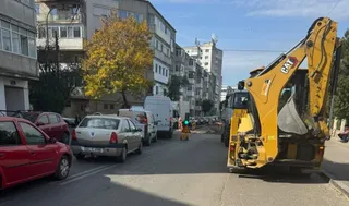 Un excavator galben este parcat pe marginea unui drum lângă mașini aliniate, în fața unor clădiri de apartamente și a unui copac cu frunze galbene.