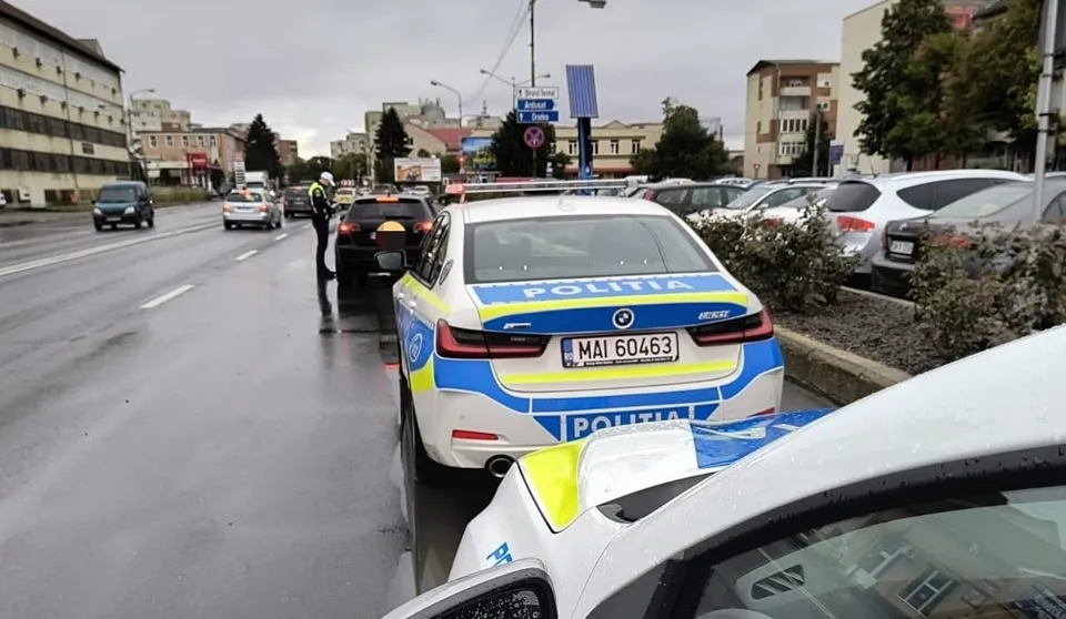 O mașină de poliție albastră și albă stă oprită pe un drum umed lângă alte vehicule parcate și în mișcare sub un cer gri.