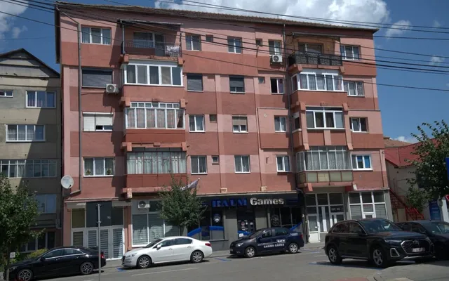 O clădire de apartamente cu fațadă roz și maro prezintă ferestre, balcoane închise și magazine la parter, cu mașini parcate în fața acesteia.