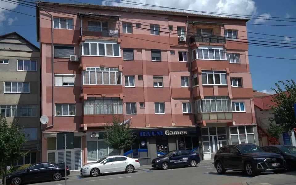 O clădire de apartamente cu fațadă roz și maro prezintă ferestre, balcoane închise și magazine la parter, cu mașini parcate în fața acesteia.