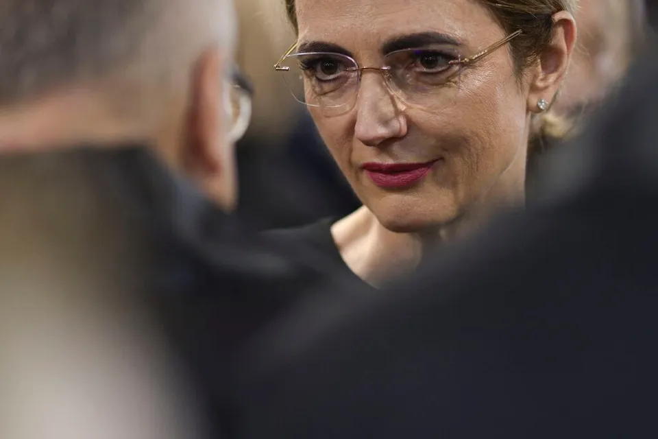 O femeie cu ochelari și ruj roșu privește spre o persoană aflată în prim-plan, cu spatele la cameră.