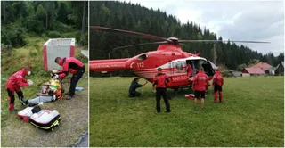 O echipă de salvatori în uniforme roșii stă lângă un elicopter roșu aterizat pe o iarbă, în timp ce alți salvatori asistă o persoană întinsă pe sol.