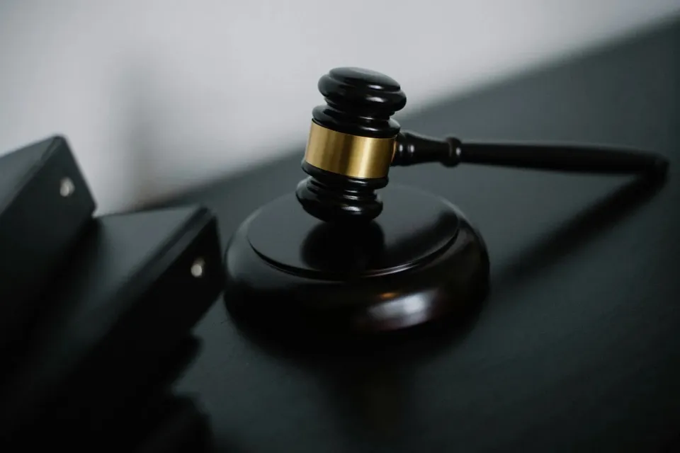 Un ciocan de judecată din lemn negru cu detalii aurii este așezat pe o bază rotundă pe o suprafață întunecată lângă niște obiecte negre.