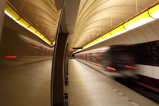 Un tren de metrou în mișcare trece printr-o stație subterană cu tavan curbat și lumini alungite pe deasupra peronului.