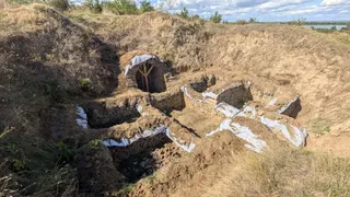 O groapă mare de săpătură arată resturi de ziduri de piatră parțial acoperite cu materiale plastice albe sub un cer înnorat.