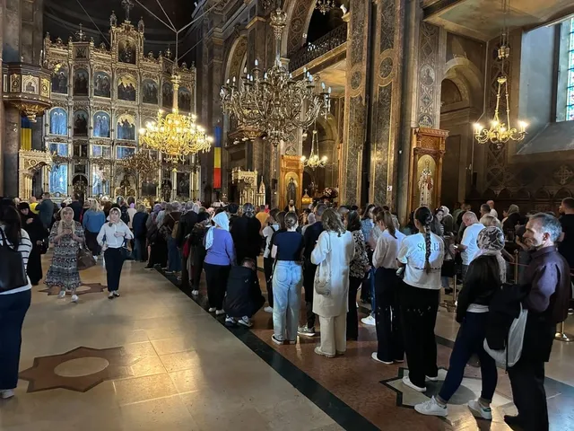 Mulți oameni stau în picioare într-o catedrală decorată cu iconostas, candelabre mari și picturi murale pe pereți.