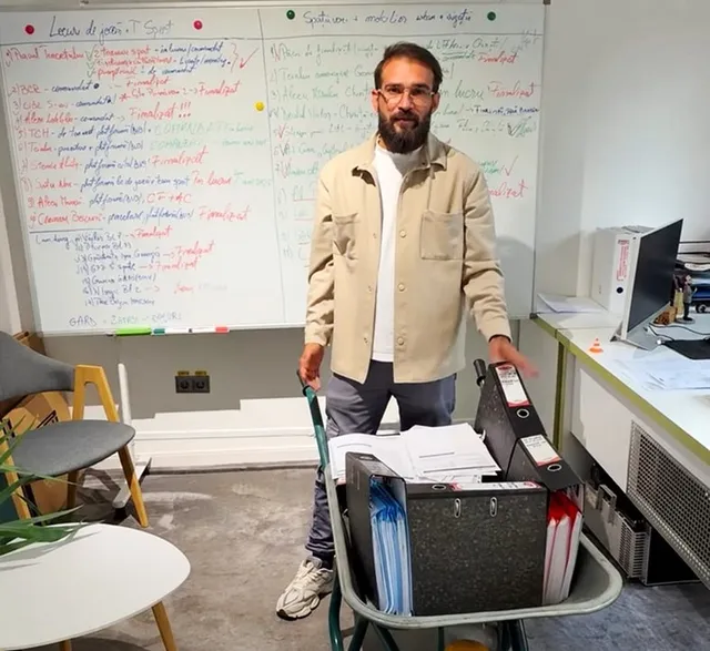 Un bărbat cu barbă și ochelari stă în picioare lângă un scaun pe care se află dosare și hârtii, în fața unui panou alb cu scrieri colorate.