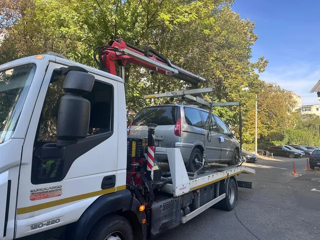 Un camion alb cu platformă transportă un autoturism gri pe un drum asfaltat lângă copaci verzi.