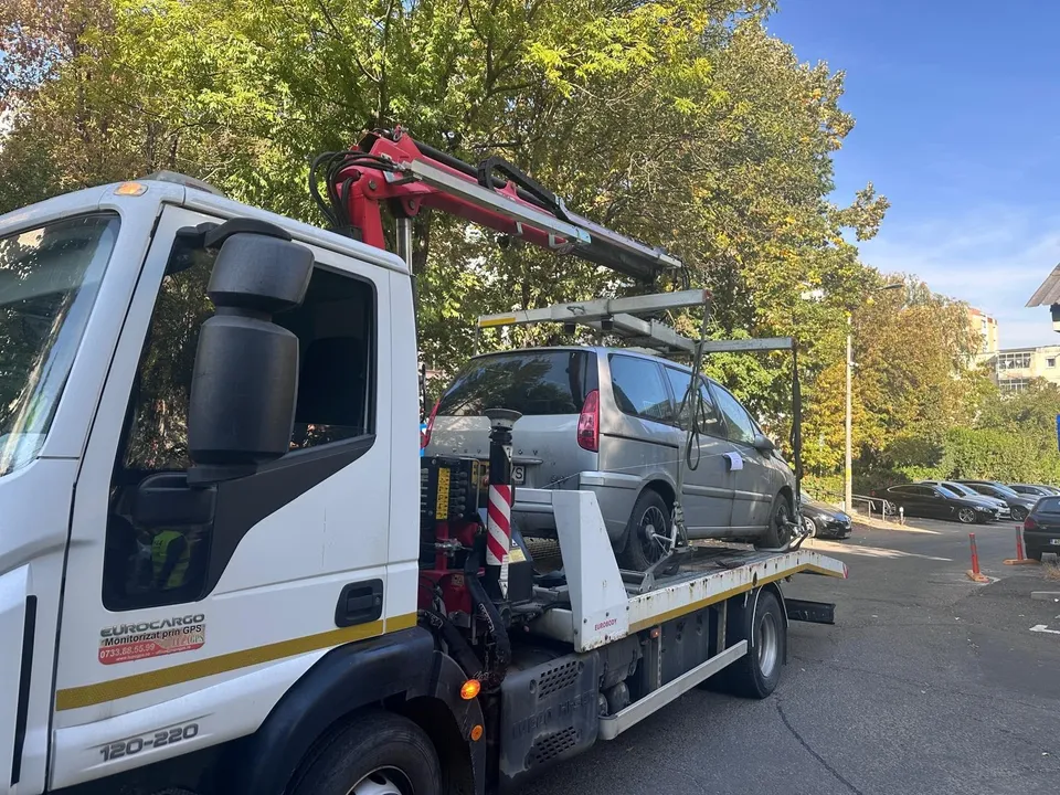 Un camion alb cu platformă transportă un autoturism gri pe un drum asfaltat lângă copaci verzi.
