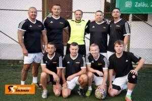 O echipă de minifotbal masculin, în uniforme negre și albe, pozând grupată pe un teren de iarbă sintetică.