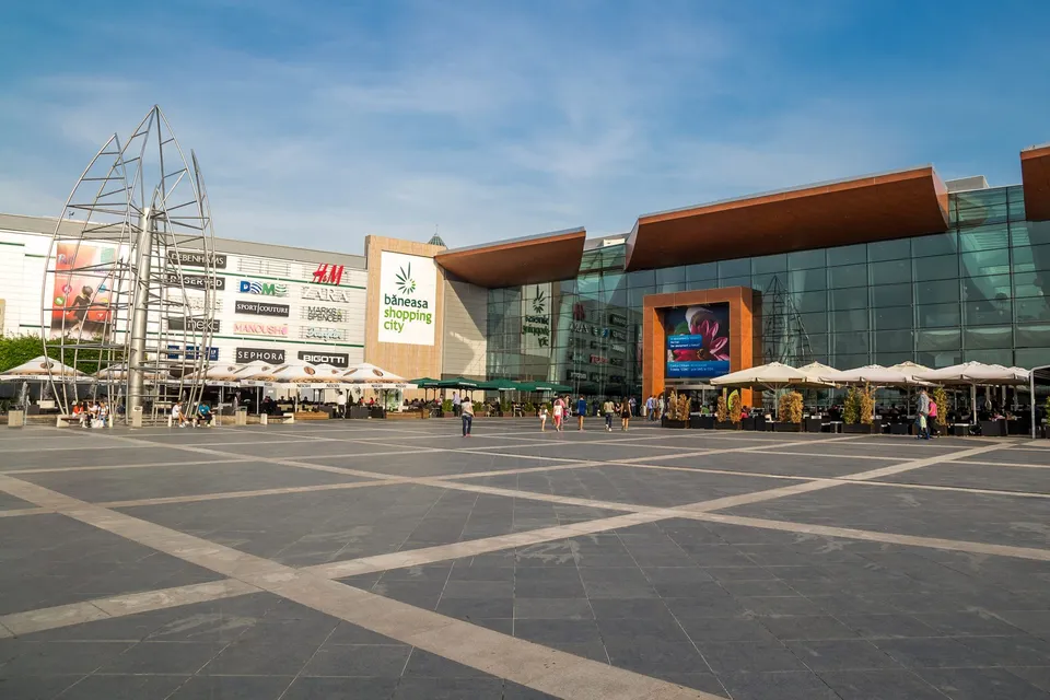 O mare piață pavată cu dale gri deschise, cu linii de împărțire albe, în fața unui complex comercial modern cu fațadă de sticlă, panouri metalice maro și o mare structură metalică de pe stânga.