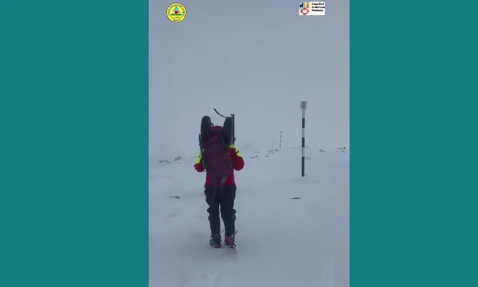 O persoană cu un rucsac mare și echipament de munte merge pe un teren acoperit de zăpadă sub un cer gri.