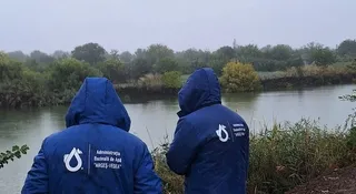 Două persoane purtând jachete albastre cu glugă și un logo alb stau cu spatele, privind spre un râu înconjurat de vegetație sub un cer gri.