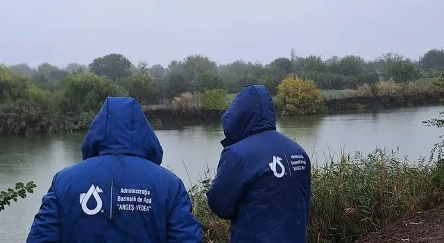 Două persoane purtând jachete albastre cu glugă și un logo alb stau cu spatele, privind spre un râu înconjurat de vegetație sub un cer gri.
